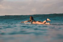Вид сбоку веселой женщины, плавающей на доске для серфинга между морской водой и голубым небом на Бали, Индонезия — стоковое фото