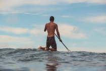 Visão traseira do remo masculino na prancha de surf entre a água do mar e o céu azul em Bali, Indonésia — Fotografia de Stock