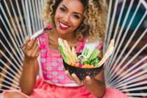 Black young woman eating fresh vegetables with dip in bowl while sitting on wicker chair — Stock Photo