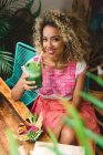 Happy black young woman holding glass of shake and sitting near wooden table with bowl of vegetables in cafe — Stock Photo