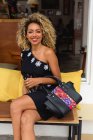 Portrait of stylish black young woman with curly hair and handbag in dress sitting on bench — Stock Photo