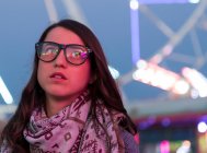 Senhora alegre em faixa e óculos perto da roda gigante destacada no parque de diversões à noite no fundo borrado — Fotografia de Stock