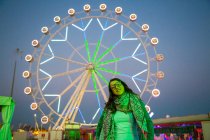 Allegro signora in fascia e occhiali vicino evidenziato ruota panoramica nel parco divertimenti in serata su sfondo sfocato — Foto stock