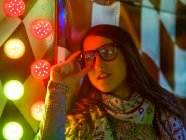 Trendy confident lady in sash and sunglasses near wall with neon lights on street at night — Stock Photo