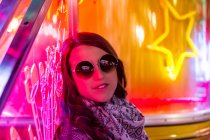 Moda señora confiada en el marco y gafas de sol cerca de la pared con luces de neón en la calle por la noche - foto de stock