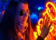 Senhora confiante na moda em faixa e óculos de sol perto da parede com luzes de néon na rua à noite — Fotografia de Stock