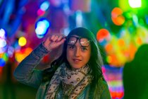 Trendy selbstbewusste Dame mit Schärpe und Sonnenbrille in der Nähe von Neonlichtern auf der Straße in der Nacht auf verschwommenem Hintergrund — Stockfoto