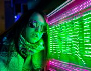Trendy signora fiduciosa in fascia e occhiali da sole vicino alla parete con luci al neon sulla strada di notte — Foto stock