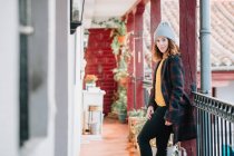 Позитивна приваблива молода жінка в теплому одязі і капелюсі, дивлячись в сторону будинку і паркану — стокове фото