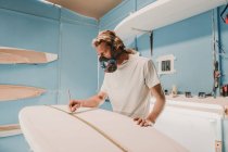 Человек в респираторе измеряет доску для серфинга в мастерской — стоковое фото