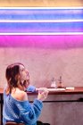 Удовлетворенная молодая женщина в трикотажном свитере держит чашку горячего напитка за столом у стены с неоновыми огнями — стоковое фото