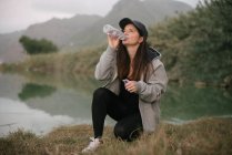 Sportswoman drinking water near lake between mountains — Stock Photo
