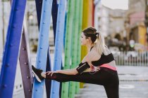 Vista laterale della donna in abbigliamento sportivo con auricolari e smartphone che allunga le gambe sulla strada — Foto stock