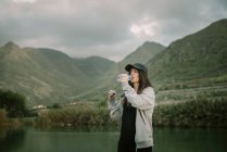 Женщина в спортивной одежде пьет воду из бутылки возле озера между высокими холмами и облачным небом — стоковое фото