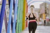 Vista posteriore della donna in abbigliamento sportivo con auricolari e smartphone in esecuzione sulla strada — Foto stock