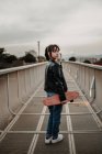 Девочка-подросток со скейтбордом стоит на металлическом мосту и смотрит через плечо — стоковое фото