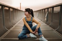 Chica sentada en monopatín en puente de metal y mirando hacia otro lado - foto de stock