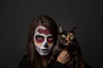 Jovem morena com maquiagem sombria segurando gato engraçado e olhando para a câmera no fundo preto — Fotografia de Stock