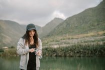 Mujer en ropa deportiva bebiendo agua cerca del lago entre montañas - foto de stock
