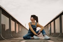 Menina sentada no skate na ponte de metal e olhando para longe — Fotografia de Stock