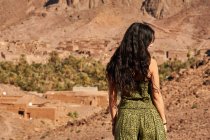 Погляд на молоду жінку з брюнетки, що стоїть між пустельними землями поблизу стародавніх споруд і пагорбів у Марракеші (Марокко). — стокове фото