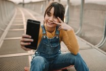 Lustiges Mädchen macht Selfie auf Smartphone, während es auf Skateboard auf Metallsteg sitzt — Stockfoto