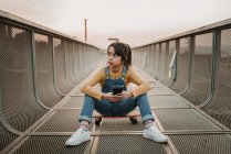 Chica con smartphone sentado en monopatín en puente de metal y mirando hacia otro lado - foto de stock