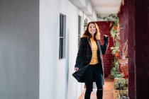 Positiv attraktive junge Frau blickt in die Kamera und geht auf Passage in Haus in der Nähe von Blumentöpfen mit Pflanzen — Stockfoto