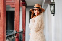 Позитивна приваблива молода жінка в теплому одязі і капелюсі дивиться на камеру і стоїть біля будинку і паркану — стокове фото