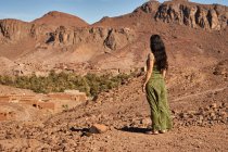 Vista posterior de la joven morena de pie entre tierras desiertas cerca de antiguas construcciones y colinas en Marrakech, Marruecos - foto de stock