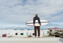 Homme debout avec planche de surf sur le toit du bâtiment et regardant la montagne — Photo de stock