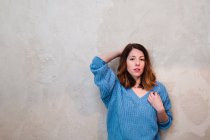 Lovely serious young woman in knitted sweater looking at camera and standing near grey wall — Stock Photo