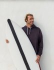Schöner erwachsener Kerl im Pullover, der mit einem Surfbrett in der Nähe der weißen Wand steht — Stockfoto