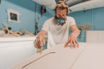 Человек в респираторе измеряет доску для серфинга в мастерской — стоковое фото