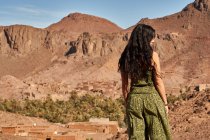 Vista posteriore di giovane signora bruna in piedi tra terre desertiche vicino a antiche costruzioni e colline a Marrakech, Marocco — Foto stock