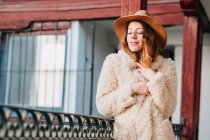 Positivo attraente giovane donna in caldo usura e cappello guardando altrove e in piedi vicino a casa e recinzione — Foto stock