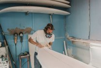 Человек в респираторе полировка доска для серфинга в мастерской — стоковое фото