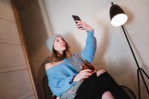 Молода жінка в трикотажному светрі з шарфом і капелюхом бере селфі на смартфон і сидить на стільці біля стіни і лампи в кімнаті — стокове фото