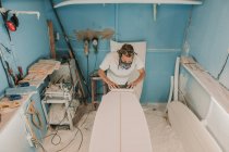 Людина в респіраторі полірує дошку для серфінгу в майстерні — стокове фото