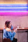 Agréable jeune femme en pull tricoté tenant tasse de boisson chaude à la table près du mur avec des néons — Photo de stock