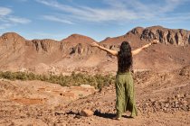 Rückansicht einer jungen brünetten Dame mit den Händen an den Seiten, die zwischen Wüstenland in der Nähe von antiken Bauten und Hügeln in Marrakesch, Marokko steht — Stockfoto