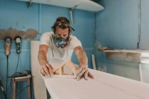 Mann mit Atemschutzmaske poliert Surfbrett in Werkstatt — Stockfoto
