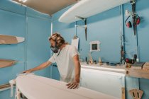 Человек в респираторе полировка доска для серфинга в мастерской — стоковое фото