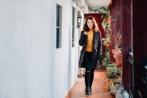 Mujer joven atractiva positiva en ropa de abrigo mirando a la cámara y pasando por casa cerca de macetas con plantas - foto de stock