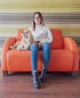 Proud stylish teen in eyeglasses sitting on armchair with dog and looking at camera in room — Stock Photo