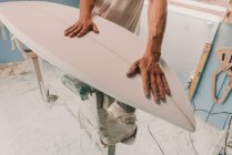Crop male holding wooden surf board placed on stand in workplace — Stock Photo