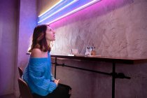 Donna positiva con coppa a tavola vicino alla parete con lampade al neon — Foto stock