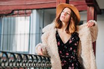 Positive attraktive junge Frau in warmer Kleidung und Hut, die wegschaut und in der Nähe von Haus und Zaun steht — Stockfoto