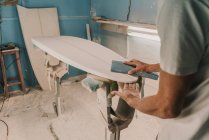 Людина в респіраторі полірує дошку для серфінгу в майстерні — стокове фото