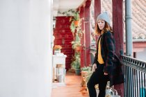 Positivo atraente jovem mulher em desgaste quente e chapéu olhando para longe e de pé perto de casa e cerca — Fotografia de Stock